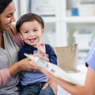 An adorable toddler boy sits in his mother's lap in his unrecognizable pediatrician's office.  He smiles and looks away.  His pediatrician holds a clipboard.