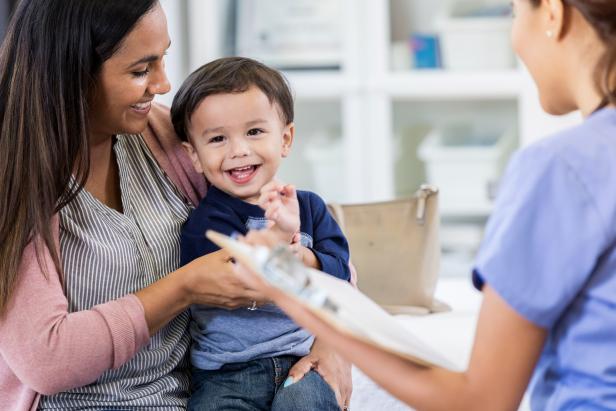 An adorable toddler boy sits in his mother's lap in his unrecognizable pediatrician's office.  He smiles and looks away.  His pediatrician holds a clipboard.