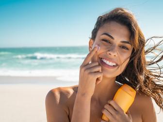 Beautiful young woman at beach applying sunscreen on face and looking at camera. Beauty latin girl enjoying summer holiday while applying suntan lotion at sea. Portrait of happy woman with healthy skin applying sunblock on cheek.