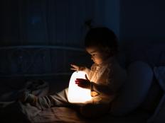 Adorable baby girl playing with bedside lamp in nursery. Happy kid sitting on bed with nightlight. Little child at home in the evening before sleep.