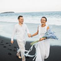 Caucasian appearance, bride in white wedding dress. Romantic feelings and positive emotions. Blue delphinium flowers bouquet. Tropical exotic destination wedding in Bali island. Black sand volcanic beach. Elopement photo session by the ocean. Beautiful scenic coastline view.