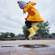 Cute and playful female child jumping in a puddle of water on the street wearing yellow rubber boots and a raincoat.