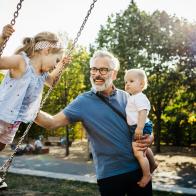 A grandfather having fun, pushing his granddaughter on the swings while holding his grandson on a day out together at the park.