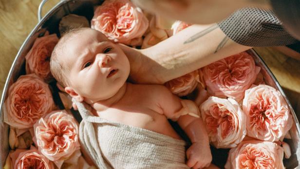Small cute neborn beautiful baby bathes in warm water with pink flowers and petals in the hands of a caring mother. The concept of baby care and aromatherapy.