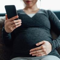 Cropped shot of pregnant woman having a video call on smartphone while touching her belly, sitting on sofa at home. Using telemedicine app on smartphone to monitor her health.