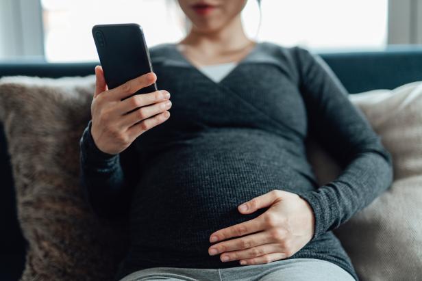 Cropped shot of pregnant woman having a video call on smartphone while touching her belly, sitting on sofa at home. Using telemedicine app on smartphone to monitor her health.
