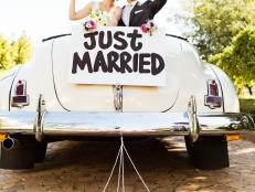 Happy bride and waving in convertible car with cans and just married sign attached to it. Vertical shot.