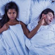 Photo of an attractive young mixed-race couple lying on their bed.  Man is snoring, keeping annoyed wife wide awake.