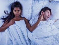 Photo of an attractive young mixed-race couple lying on their bed.  Man is snoring, keeping annoyed wife wide awake.