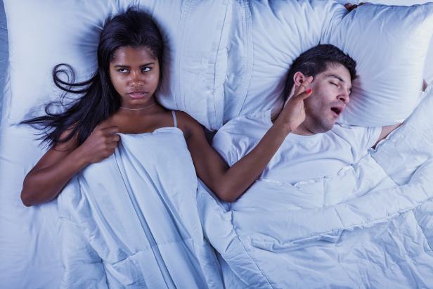 Photo of an attractive young mixed-race couple lying on their bed.  Man is snoring, keeping annoyed wife wide awake.