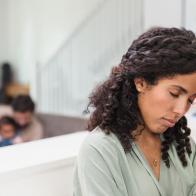 An exhausted mid adult woman rubs her head indicating a bad headache. Her husband is in the background caring for their daughter.