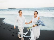 Caucasian appearance, bride in white wedding dress. Romantic feelings and positive emotions. Blue delphinium flowers bouquet. Tropical exotic destination wedding in Bali island. Black sand volcanic beach. Elopement photo session by the ocean. Beautiful scenic coastline view.