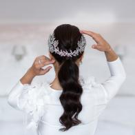 Fiancee with dark long hair turned her back to the camera. Hairstyle decorated with silver floral barrette looks amazing on the light background and pure morning light. Bride touches her head tenderly