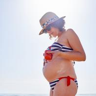 Pregnant woman putting on sunscreen on the beach with the sea in the background, she is wearing sunglasses and a hat