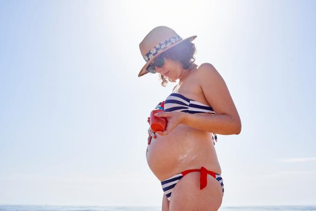 Pregnant woman putting on sunscreen on the beach with the sea in the background, she is wearing sunglasses and a hat
