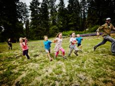 Camp counselor playing tag with group of laughing young kids in grass field at summer camp