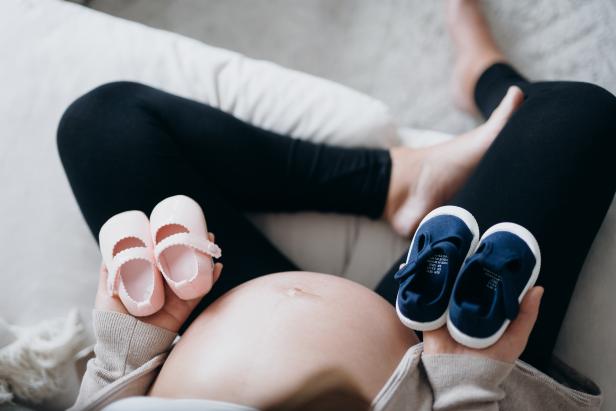Overhead view of Asian pregnant woman holding a pair of blue and pink baby shoes in front of her belly. Expecting a new life, mother-to-be, gender reveal concept