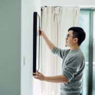 Young Asian man holding a picture frame and hanging the painting on the wall decorating in his newly refurbished apartment