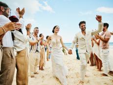 Wide shot of bride and groom walking down aisle after wedding ceremony on tropical beach while friends and family celebrate