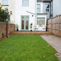 A general exterior view of a back garden patio area with wood decking, potted plants, Dragon palm tree, metal table and two chairs pale pastel sage green patio doors, window and drainpipe, grass lawn, crazy paving path, timber fencing looking towards the rear of a house