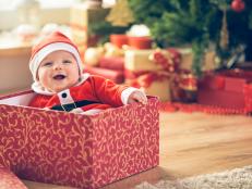 Cute little baby boy in Santa hat sitting inside of the red gift box next to the Christmas tree.