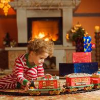 Girl, sitting by fireplace among Christmas decoration. She wearing striped pajama, she has curly hair. Girl playing with christmas train .