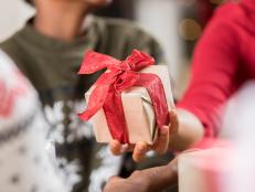 An unrecognizable woman gives a gift to a family member on Christmas Day. Close up is on the present. The present is wrapped in gold wrapping paper and tied with a red bow.