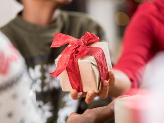 An unrecognizable woman gives a gift to a family member on Christmas Day. Close up is on the present. The present is wrapped in gold wrapping paper and tied with a red bow.