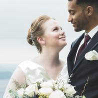 In this closeup, a beautiful bride stands outdoors with her groom and smiles up at him.  He looks down and smiles at her.  There is a scenic background.