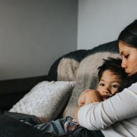 A mother and son sit on a sofa in a house environment. She tenderly embraces him and kisses his head as he snuggles into her. Wall provides a space for copy.