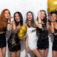 Young girlfriends celebrating bride night. Bride is wearing a veil and a T-shirt with the inscription ''BRIDE'', bridesmaids are wearing T-shirts with the inscription ''Squad''.  They are having fun and blowing confetti from hands.  Room is decorated with star and heart shaped balloons.