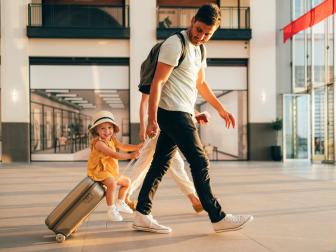 Cheerful husband and his anonymous wife walking with their little girl sitting on luggage at the airport.