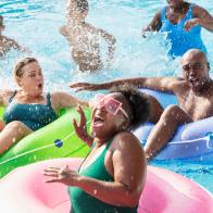 A multiracial group of six people, two families, having fun at a water park on the lazy river. The three parents are floating in inflatable rings. The three children are standing waist deep in the water behind them. The African-American woman in the foreground is laughing as she gets splashed.