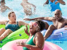 A multiracial group of six people, two families, having fun at a water park on the lazy river. The three parents are floating in inflatable rings. The three children are standing waist deep in the water behind them. The African-American woman in the foreground is laughing as she gets splashed.