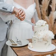 Bride and groom cutting stylish wedding cake at wedding in outdoor. Wedding couple holding knife and cutting together wedding cake decorated with flowers