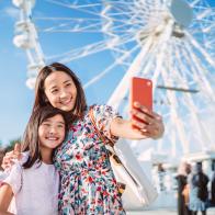 Pretty young Asian mom taking selfie with her lovely daughter against a ferris wheel when enjoying spending time together in theme park.