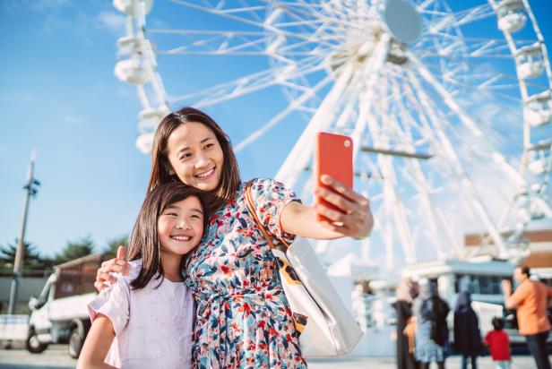 Pretty young Asian mom taking selfie with her lovely daughter against a ferris wheel when enjoying spending time together in theme park.
