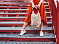 Unrecognizable young woman in red dress standing on steel grate stairs holding shopping net bag with fruits and vegetables.