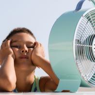 Overheated little child is sitting in front of electric fan trying to cool down.