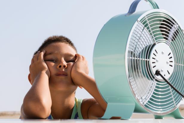 Overheated little child is sitting in front of electric fan trying to cool down.