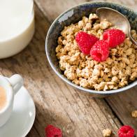 "A high angle close upshot  of a bowl full of crunchy granola with ripe red raspberries, a container of milk and a small cup with freshly brewed, foamy expresso. Shot on a grungy old wooden table."