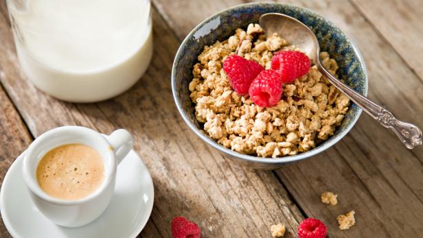 "A high angle close upshot  of a bowl full of crunchy granola with ripe red raspberries, a container of milk and a small cup with freshly brewed, foamy expresso. Shot on a grungy old wooden table."