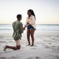 Young African man kneeling in the sand and proposing to his smiling girlfiend on a beach at dusk