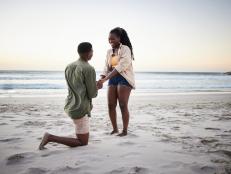Young African man kneeling in the sand and proposing to his smiling girlfiend on a beach at dusk