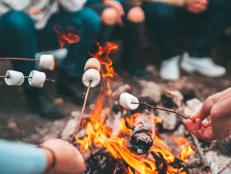 Closeup shot of friends roasting marshmallows over the bonfire with wooden skewer sticks. Autumn camping and camp food preparing outdoors, sitting around warm fire