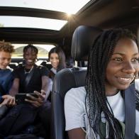 Happy teenage girl driving friends riding in back seat, using smart phone in car with sunroof