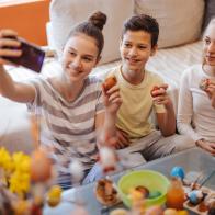 Three children, brother and two sisters sitting on floor in living room at home, holding colorful Easter eggs and taking selfie on mobile phone