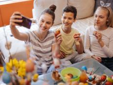 Three children, brother and two sisters sitting on floor in living room at home, holding colorful Easter eggs and taking selfie on mobile phone