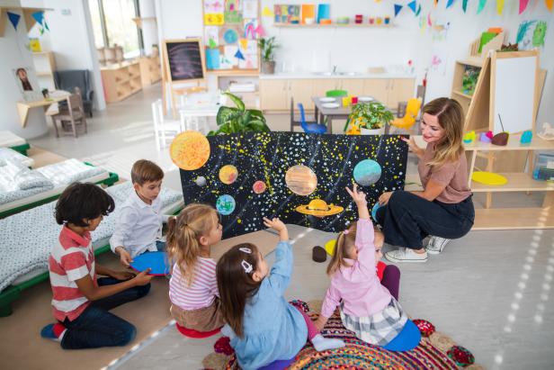 Multiracial group of small children with teacher sitting on floor and learning about space together at kindergarten.