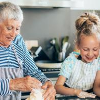 Three generation women kneading cookie dough in kitchen. Happy grandmother, mother and little girl in aprons with messy dough hands making biscuits on kitchen counter and playing with dough. Using digital tablet looking for new recipe, or watching cooking tutorial or making video vlog;